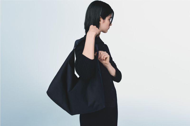 A woman with dark hair carrying a large black hobo bag. She is wearing a black cardigan and standing against a gradient blue-white background.