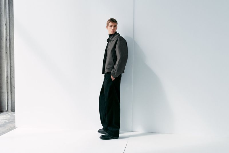 A person stands in a minimalist style, wearing a jacket, turtleneck, and trousers, with modern concrete and white walls as a backdrop.