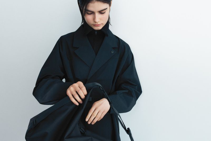 A young woman in a dark coat stands in front of a white wall, looking into a dark bag she holds open with both hands.