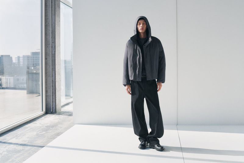 Fashion shot of a slender Asian male model posing in a hooded puffer jacket, loose black trousers, and shoes in an indoor studio setting.