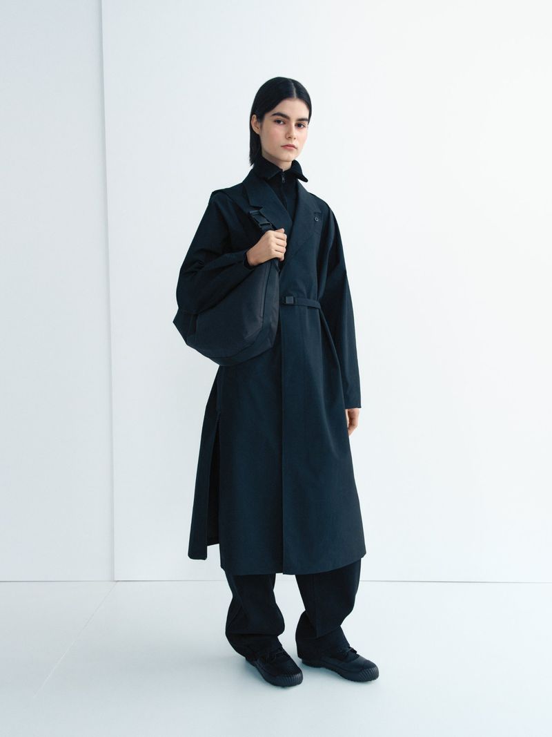 A female fashion model poses in a long black coat against a white background, carrying a black bag. Minimalist design and studio lighting.