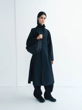 A female fashion model poses in a long black coat against a white background, carrying a black bag. Minimalist design and studio lighting.