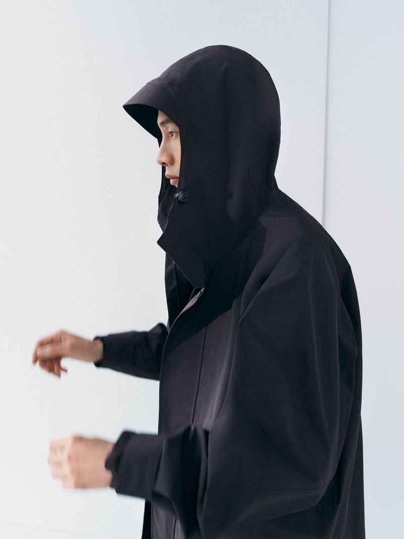 Profile view of an adult male wearing a dark hooded coat against a plain white background in a studio setting, showcasing fashion apparel.