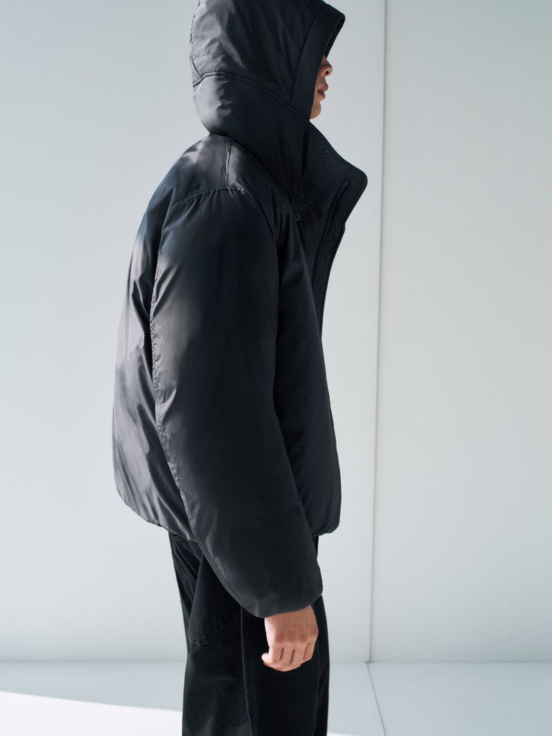 Person wearing a black hooded puffer jacket in a studio setting. The jacket is puffy and the background is a plain white wall.
