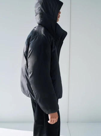 Person wearing a black hooded puffer jacket in a studio setting. The jacket is puffy and the background is a plain white wall.
