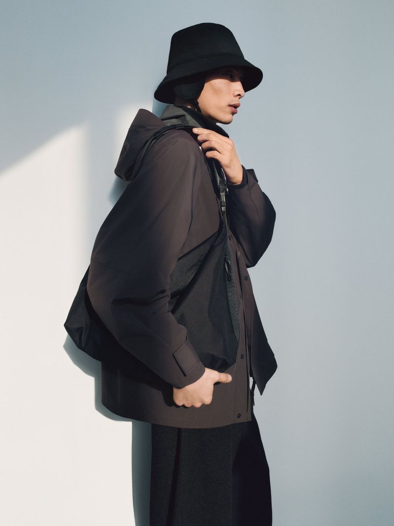 Fashion portrait of an Asian male model in a black bucket hat, jacket, and pants against a white wall with natural lighting.