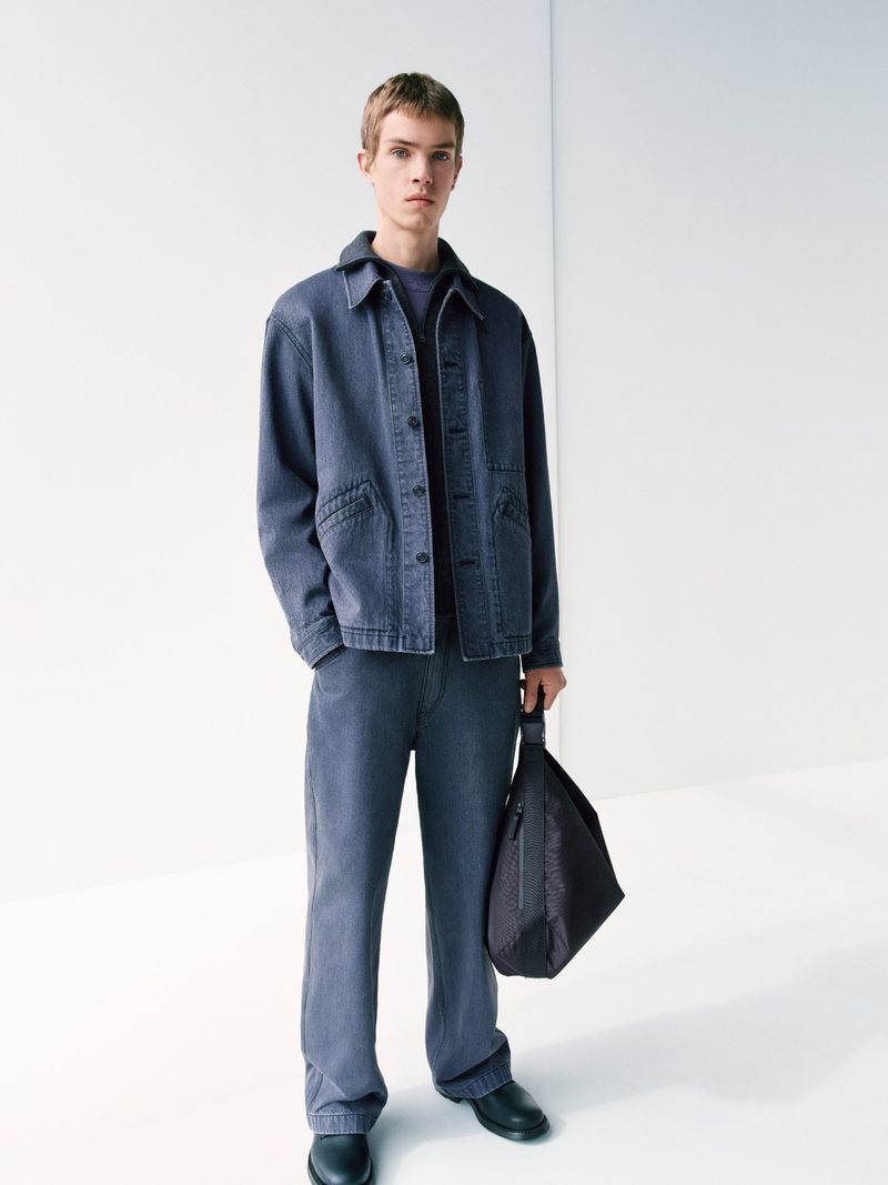 Full-length shot of a young adult male standing in a blue denim jacket and matching pants, holding a black bag against a white backdrop.