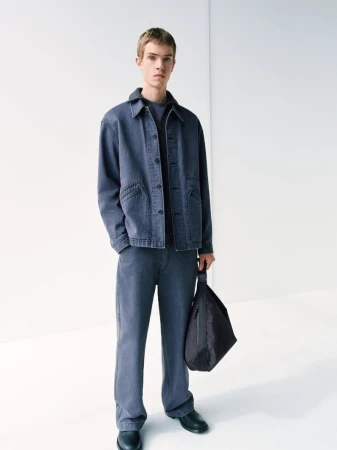 Full-length shot of a young adult male standing in a blue denim jacket and matching pants, holding a black bag against a white backdrop.