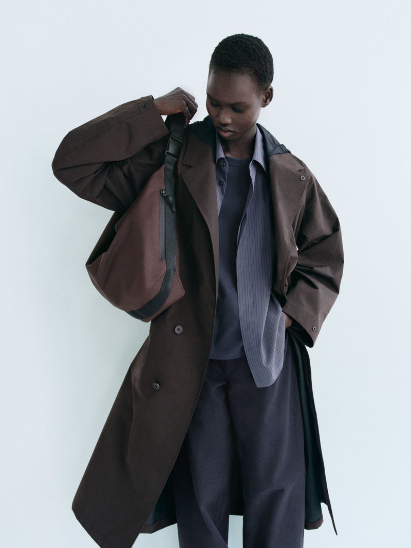 Dark-skinned adult posing with a brown overcoat and shoulder bag in a studio setting. Minimalist fashion with neutral colors.