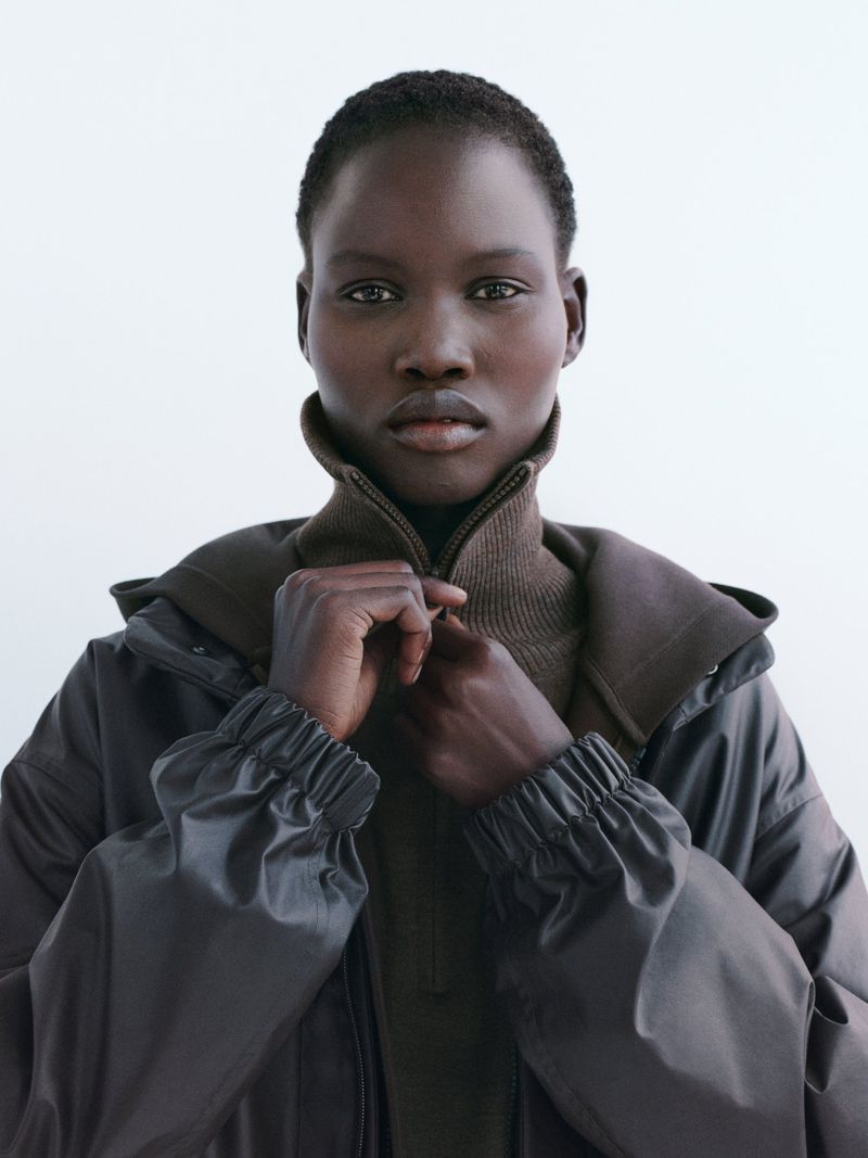 Portrait of a Black woman in a brown jacket and turtleneck, eyes forward, against a clean white background in a fashion shot.