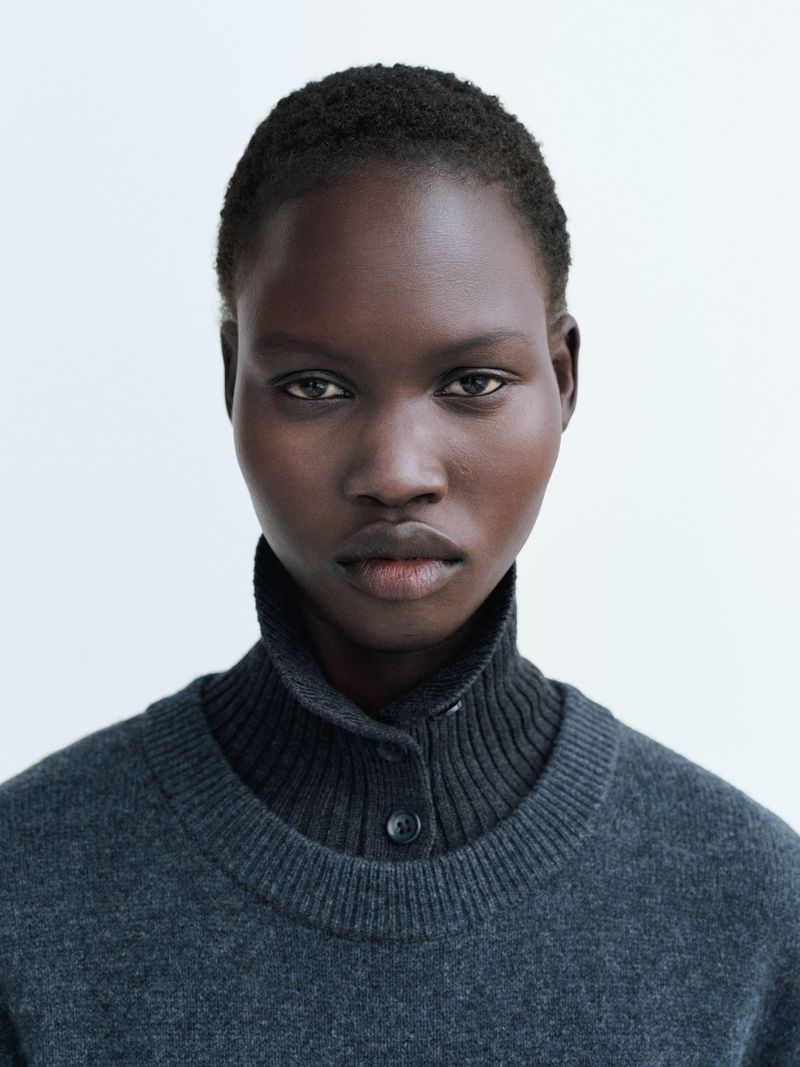 Portrait of a Black woman with short hair wearing a gray sweater and dark turtleneck against a plain white backdrop in a studio setting.
