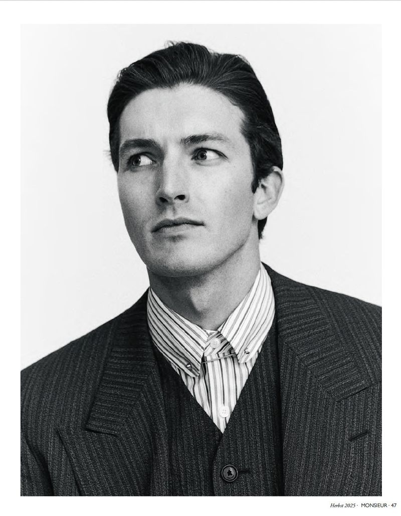 Black-and-white portrait of a man in a pinstripe suit looking to the left. He wears a striped collared shirt and vest.