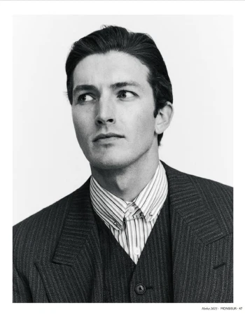 Black-and-white portrait of a man in a pinstripe suit looking to the left. He wears a striped collared shirt and vest.