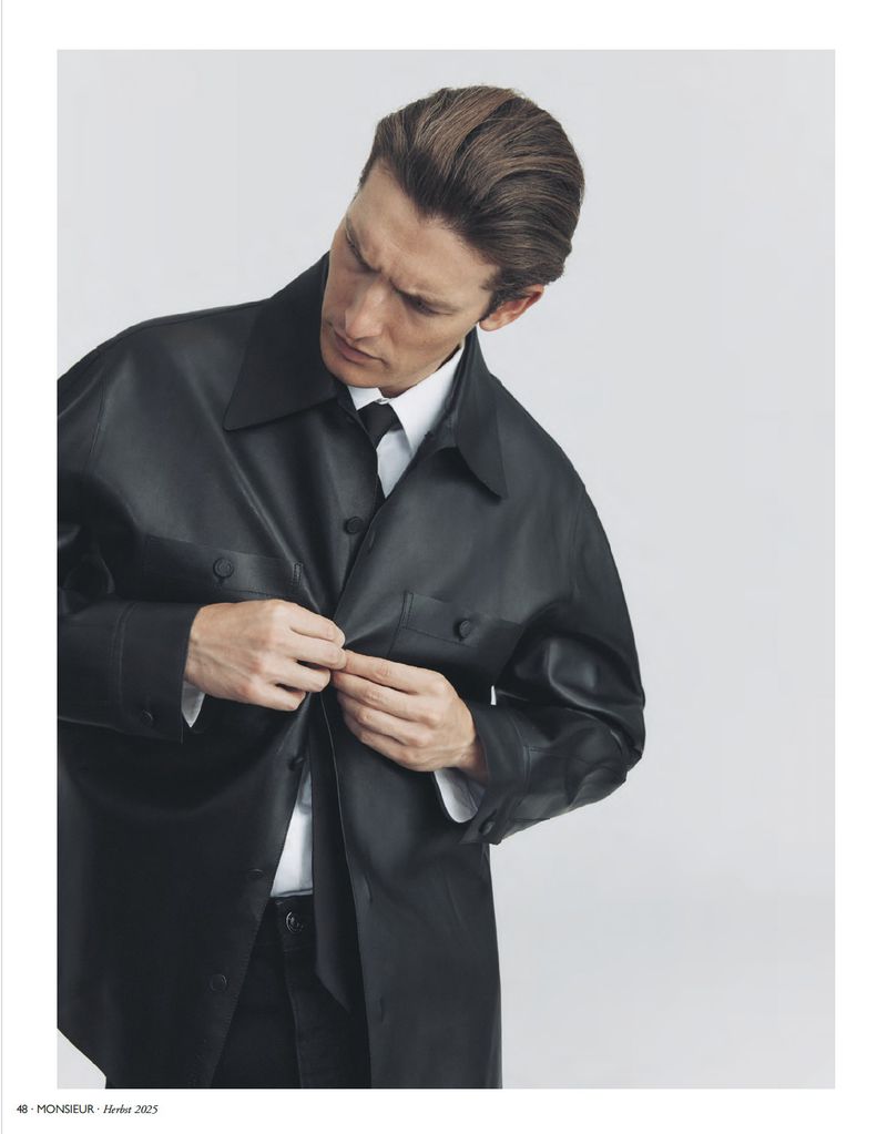 Portrait of a fair-skinned adult male buttoning a black, oversized leather shirt over a white dress shirt and black tie in a studio setting.