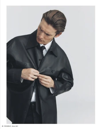 Portrait of a fair-skinned adult male buttoning a black, oversized leather shirt over a white dress shirt and black tie in a studio setting.