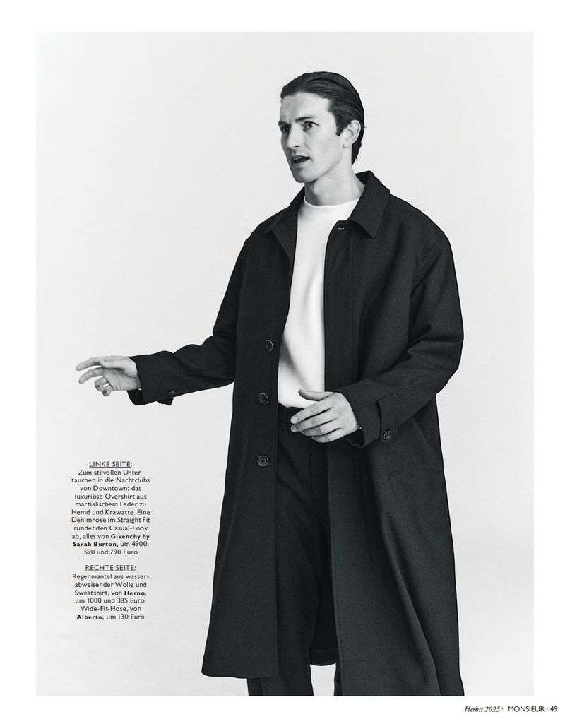 Male fashion model poses in a black overcoat and white shirt against a white backdrop in a studio setting, looking off camera.