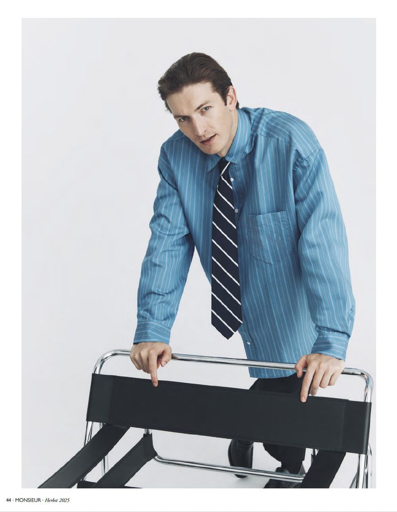 Portrait of a man in a blue striped shirt and tie, leaning on a black folding luggage rack against a plain white backdrop in a studio.