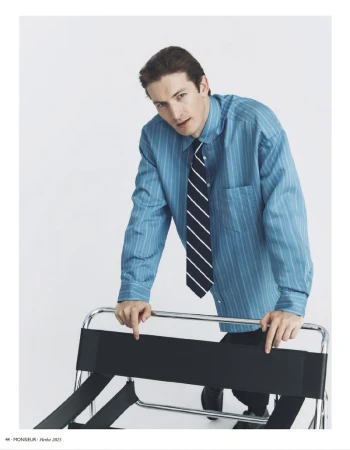 Portrait of a man in a blue striped shirt and tie, leaning on a black folding luggage rack against a plain white backdrop in a studio.