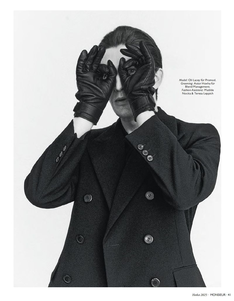 Monochrome portrait of a stylish adult male in an overcoat making binoculars with gloved hands against a bright neutral background.