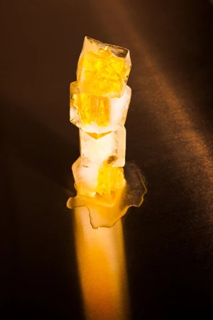 Stacked ice cubes with golden liquid inside melting under a focused light on a dark background, studio food photography.