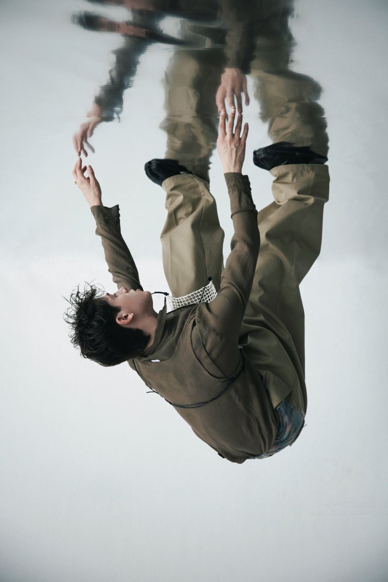 A young adult man wearing a khaki-colored jumpsuit is floating upside down, with a wavy reflection above him against a white backdrop.