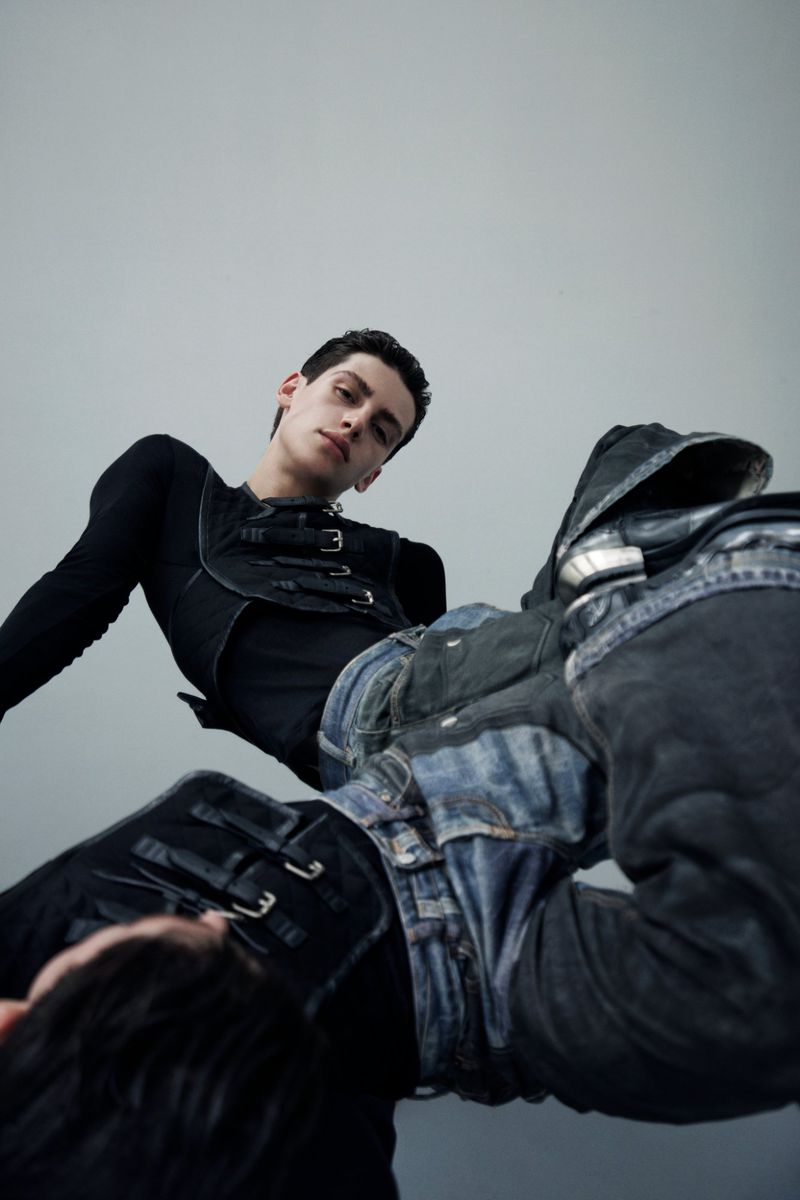 Low-angle shot of two young adult men in fashionable black tops and jeans, lying down against a plain grey backdrop in a photo studio.