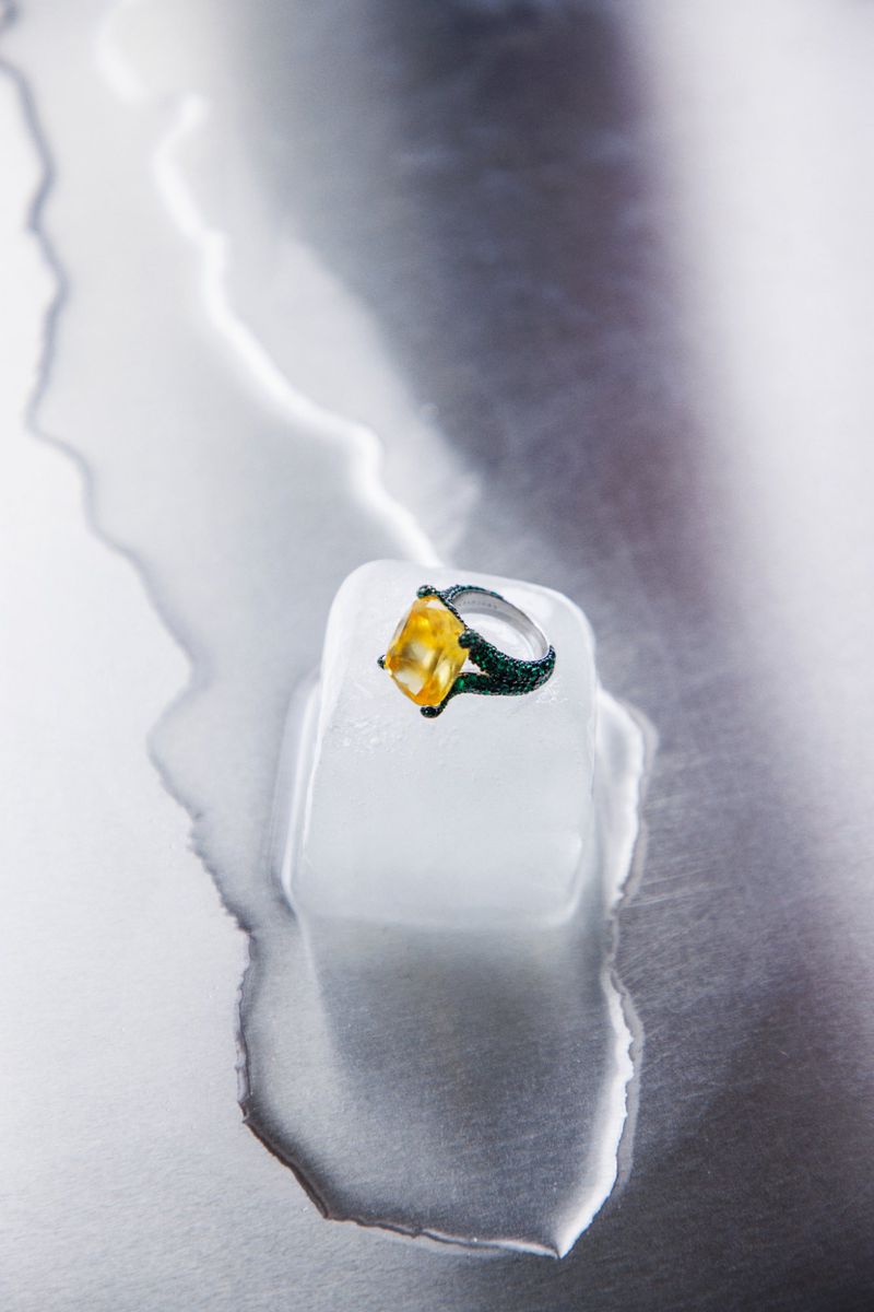 Close-up of a gemstone ring with a yellow stone sitting on a block of ice with melting ice around it on a silver background.
