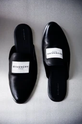 Top-down view of black leather Givenchy slip-on mule loafers with white logo patches on a neutral grey background.