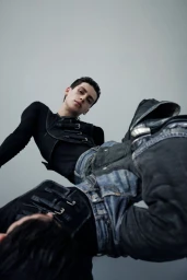 Low angle view of a young man wearing a black strapped harness vest and distressed denim jeans against a grey studio backdrop.