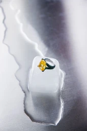 A yellow faceted gemstone ring with a green band resting on a melting ice cube over a reflective metal surface.
