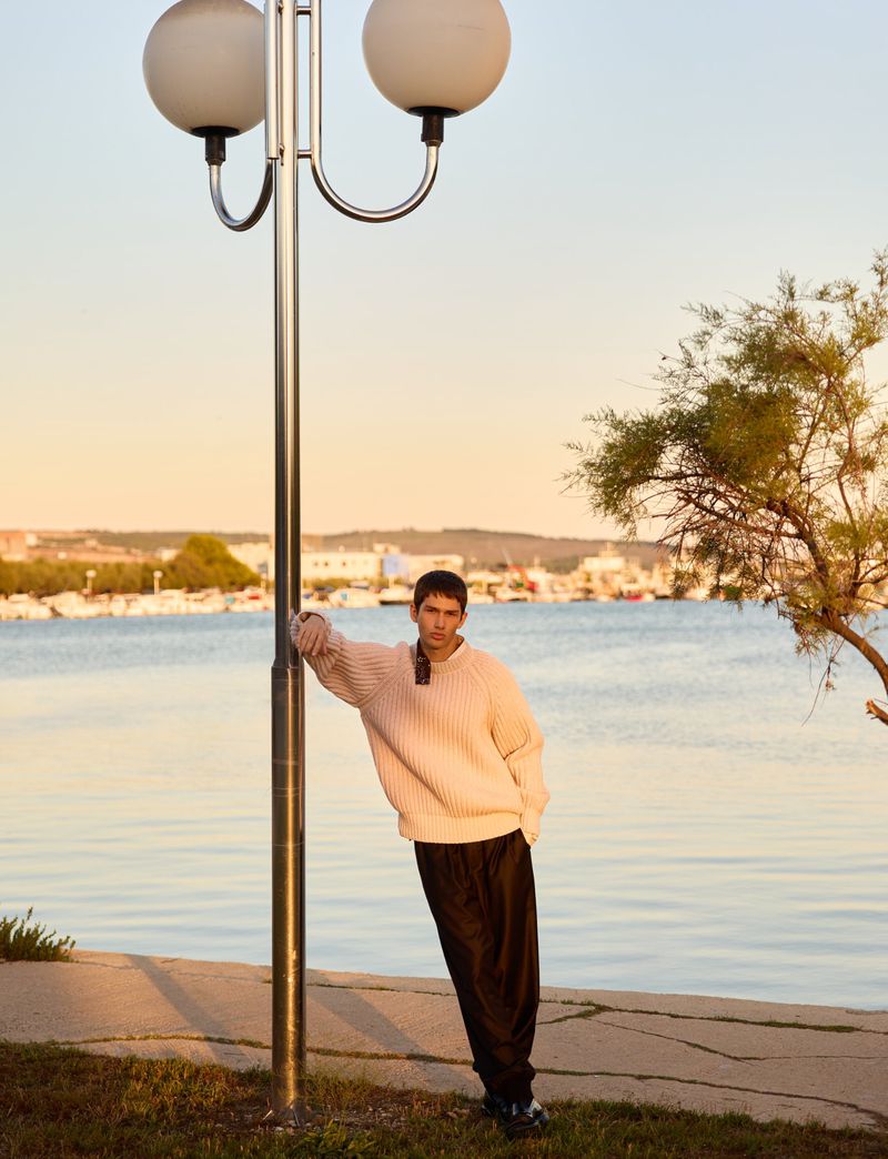 Adult man posing by a lamp post, wearing a sweater and pants, at the beach during sunset with blue ocean and docked boats.