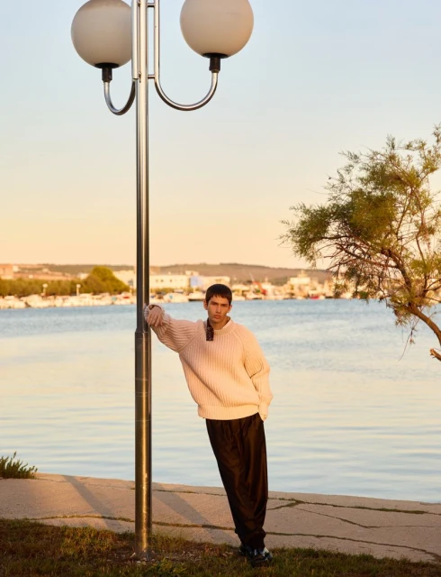 Adult man posing by a lamp post, wearing a sweater and pants, at the beach during sunset with blue ocean and docked boats.