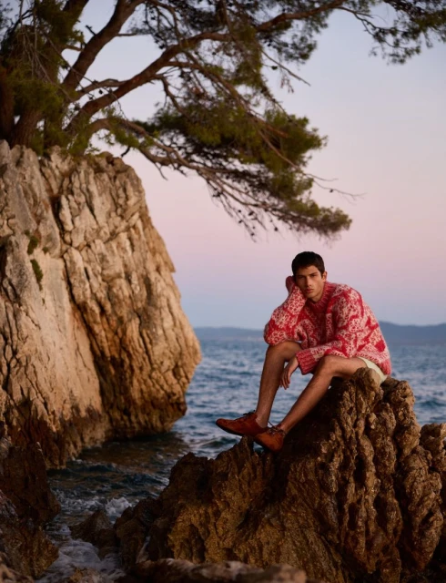 A young adult male sits on rocky outcrop by the ocean wearing a red patterned sweater, light shorts, and brown leather shoes outdoors.