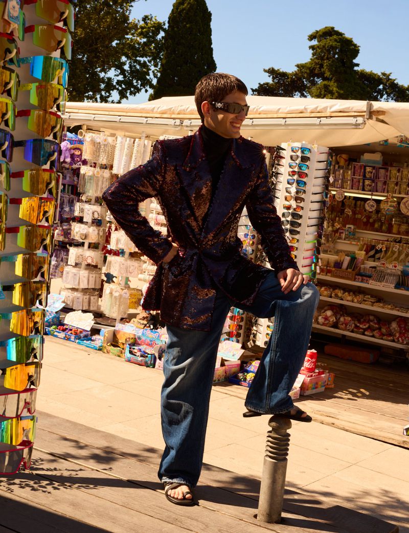 Man in sunglasses and sequin jacket poses next to sunglasses vendor on city street. He wears jeans and sandals on a sunny day.