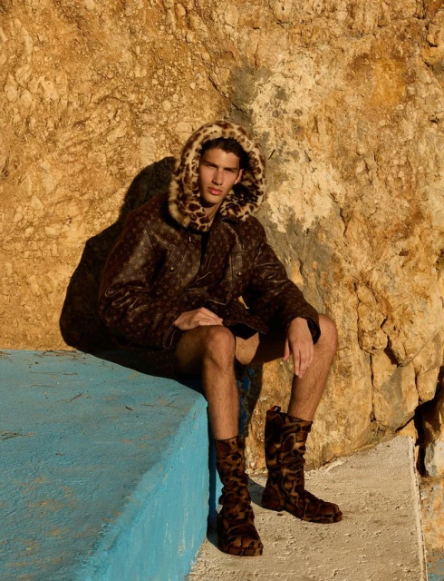Male fashion model in patterned coat with fur hood, shorts, and boots sitting on a blue ledge in front of a textured rock wall.