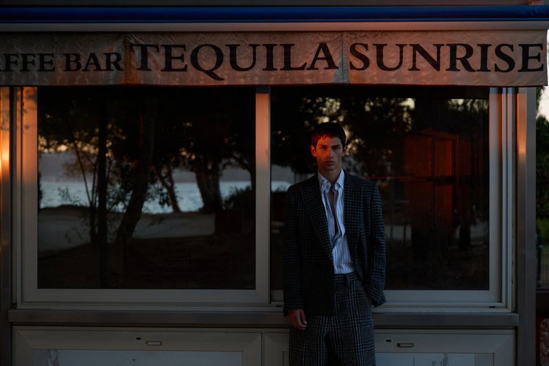 A stylish adult male poses in front of a beachside bar with a "Tequila Sunrise" sign at dusk, wearing a blazer and slacks suit.