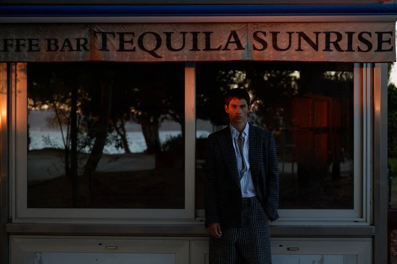 A stylish adult male poses in front of a beachside bar with a "Tequila Sunrise" sign at dusk, wearing a blazer and slacks suit.