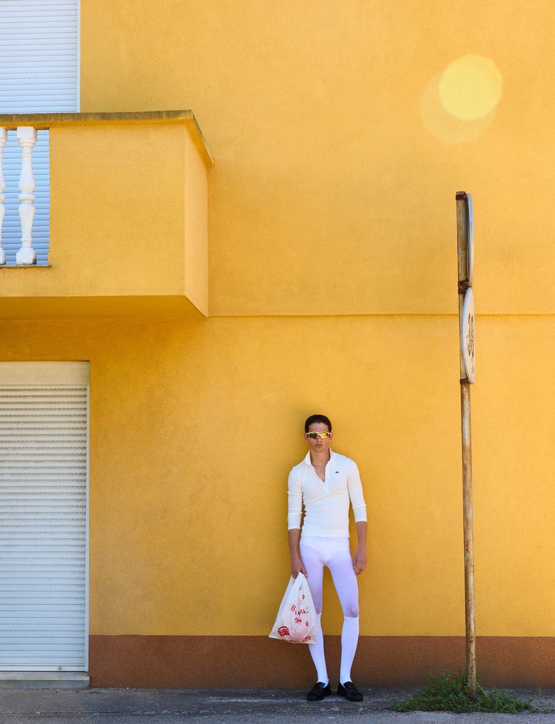 Adult male in white outfit standing against a yellow building with a plastic bag, showcasing bright outdoor fashion and minimalist urban scene.
