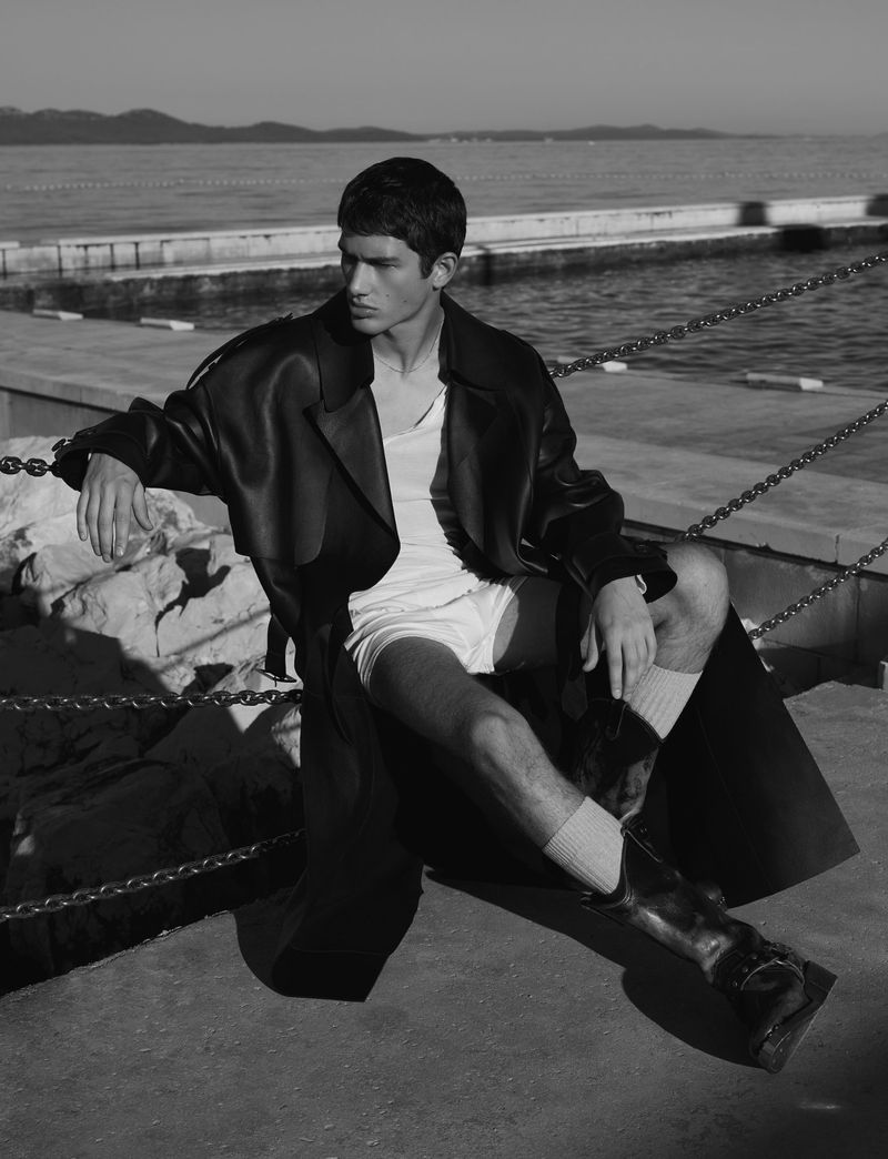 Black and white portrait of a young adult male model in a coat, tank top, shorts, boots, and socks sitting near a waterfront.