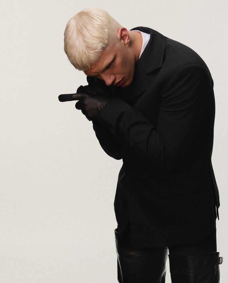 Man in black suit poses with a hand gesture resembling a gun, wearing black gloves and boots against a plain white background.