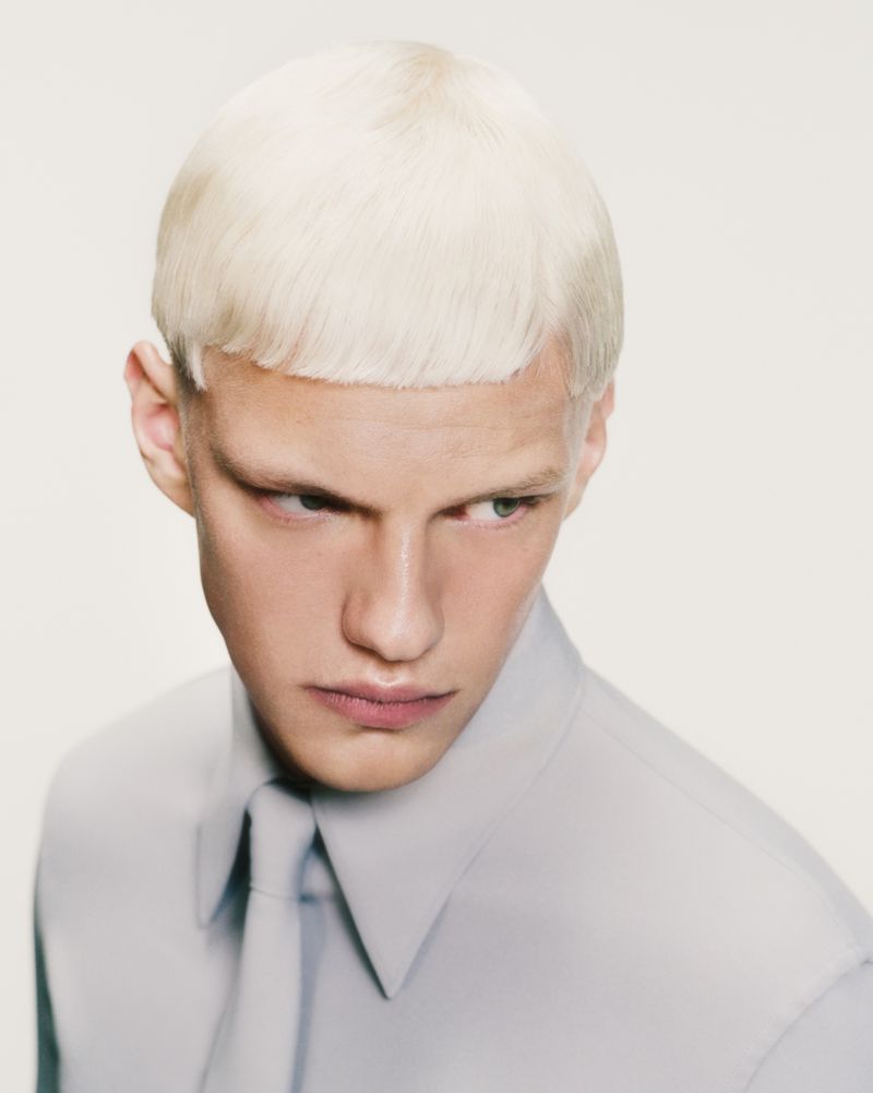 Close-up of a fair-skinned adult male with a bowl cut and light blonde hair wearing a button-down shirt and tie, gazing intently toward the side.