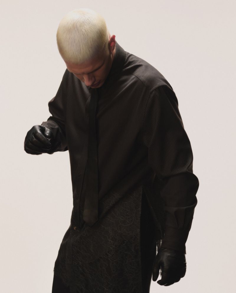 Blonde male model wearing a dark collared shirt and black leather gloves looking down against a neutral studio background.