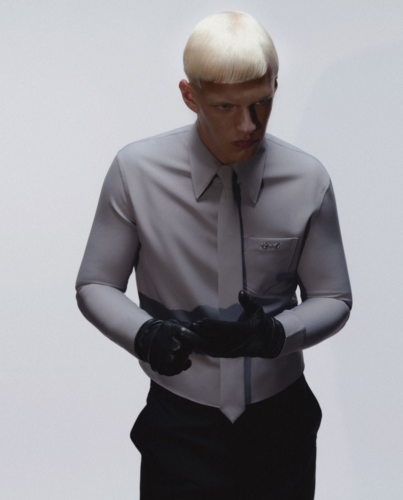 Blonde male model in gray shirt and tie, wearing black gloves, poses against a plain white background in a fashion editorial.