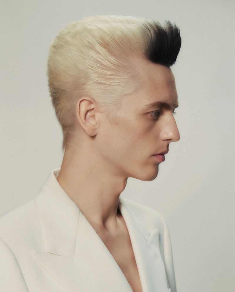 Profile view of a person with a two-toned pompadour hairstyle wearing a light-colored suit against a neutral background.