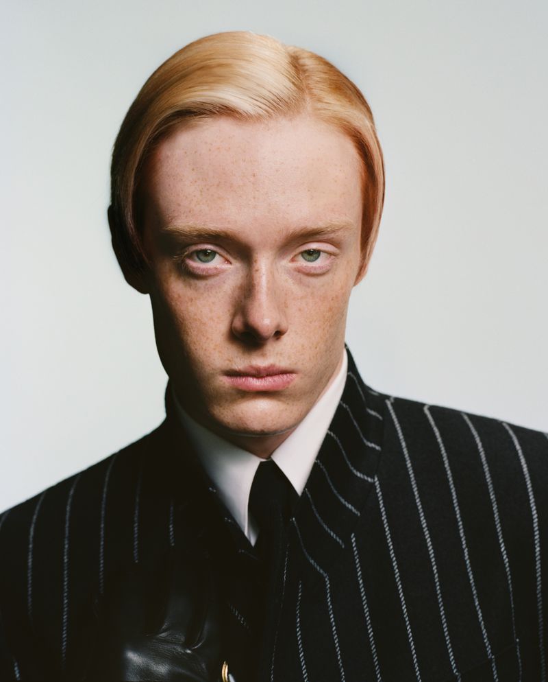 Portrait of a young adult male with fair skin and light blonde hair. He is wearing a pinstripe suit, white collared shirt and dark tie.