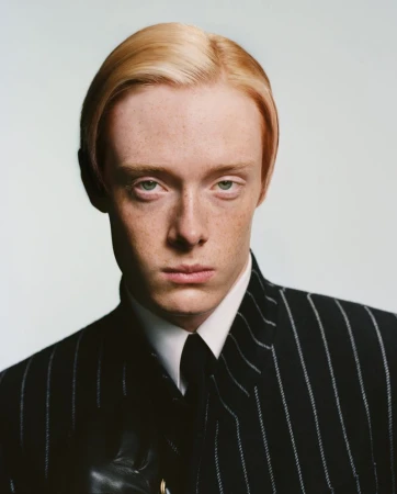 Portrait of a young adult male with fair skin and light blonde hair. He is wearing a pinstripe suit, white collared shirt and dark tie.
