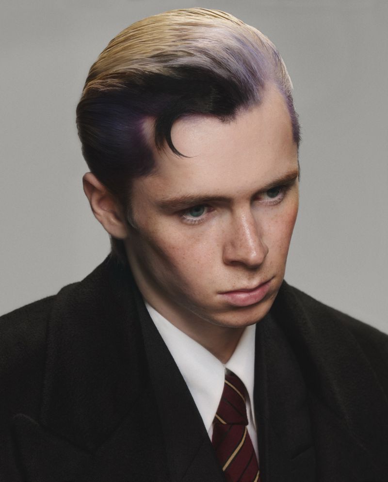 Portrait of a young adult male with dyed blonde and purple hair, wearing a dark wool coat, white shirt, and striped tie against a plain background.
