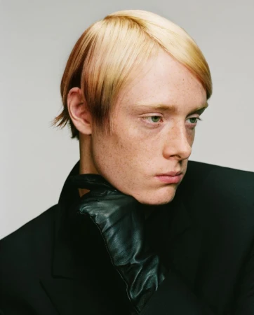 Portrait of a man with blonde styled hair wearing a black jacket and a black leather glove resting against his chin.