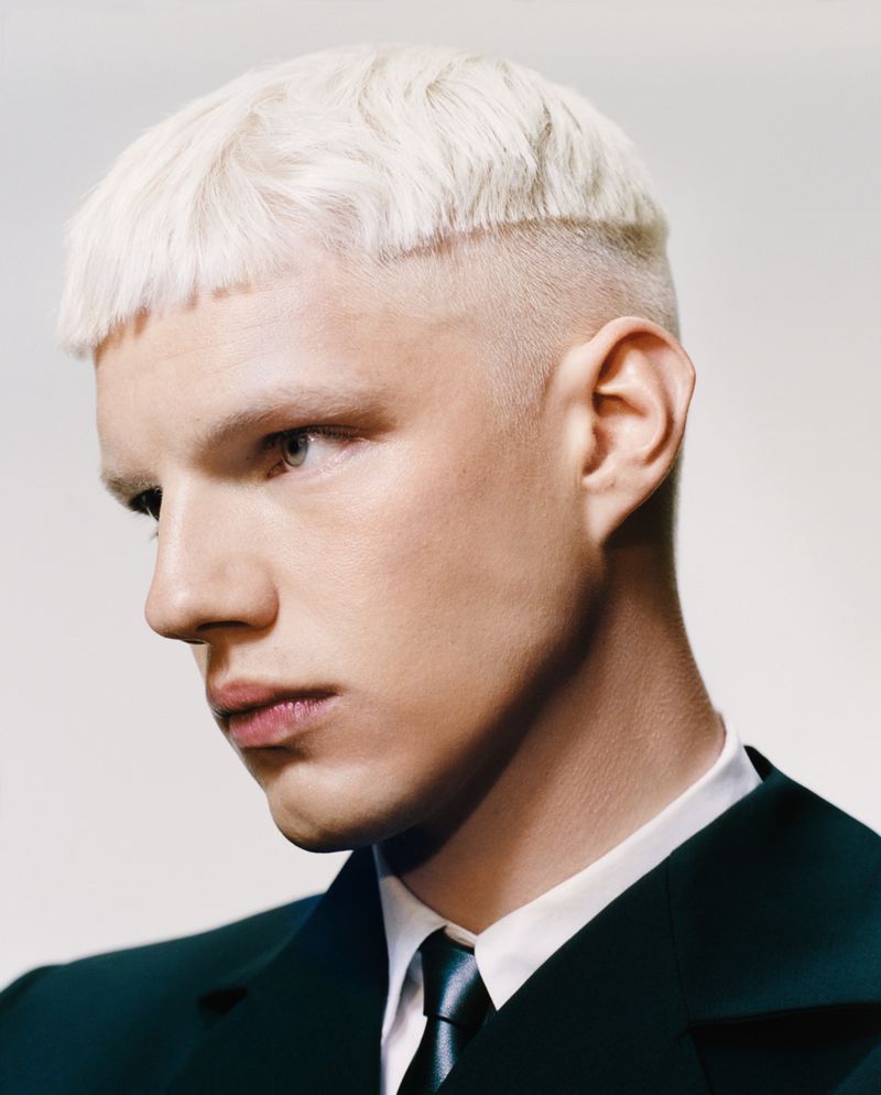 Portrait of a light-skinned adult male with a buzz cut, bleached blond hair, wearing a dark jacket and light tie in a light studio setting.
