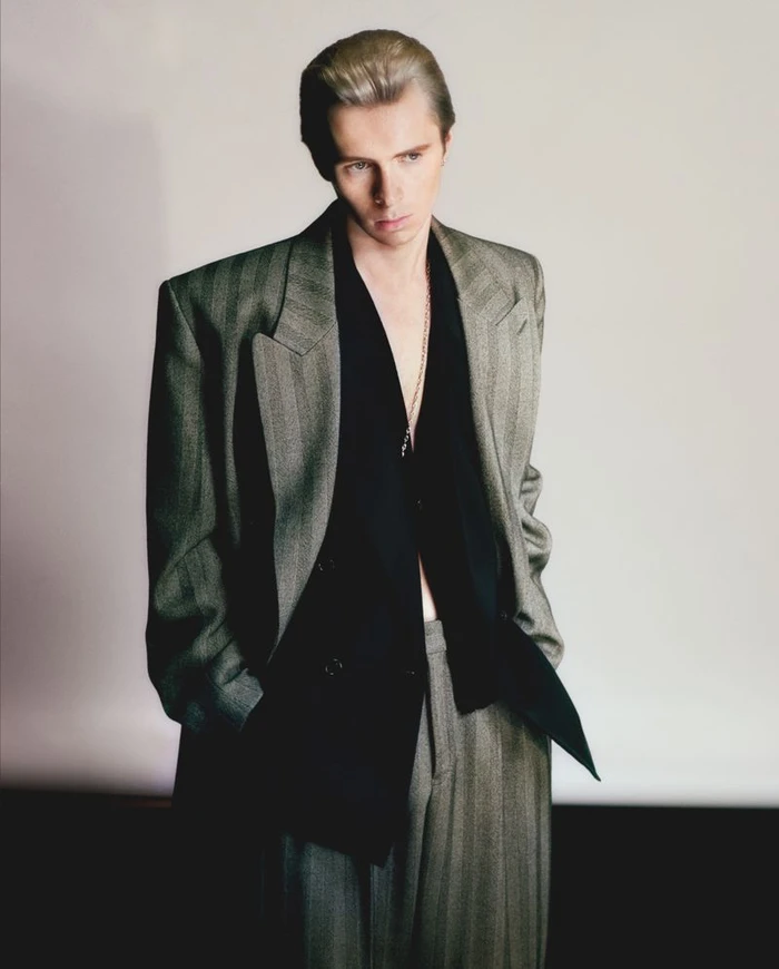 Man wearing a structured grey pinstripe blazer and trousers with a black shirt against a plain white background.